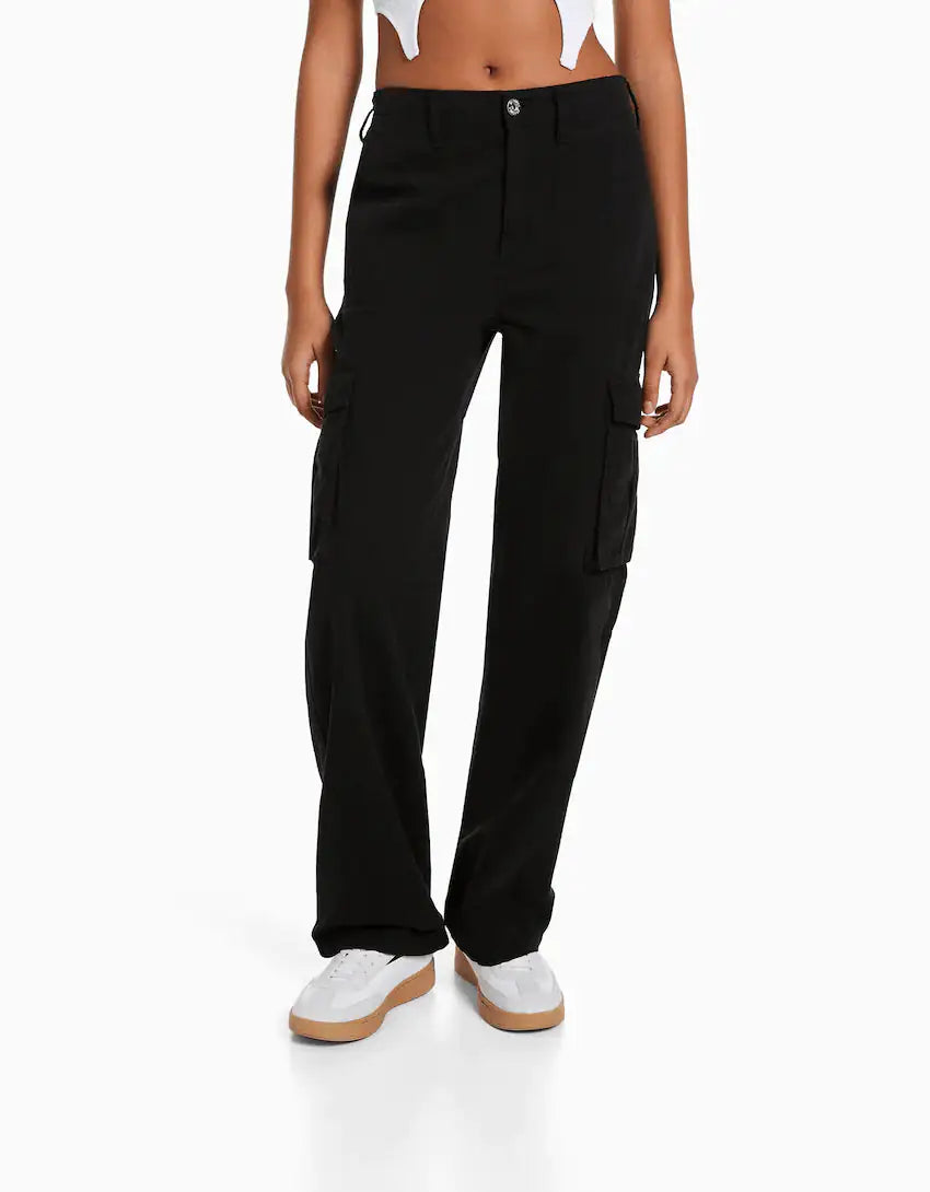 Front view of a woman wearing high-waisted black cargo trousers with flap side pockets, styled with a black crop top and black sneakers.