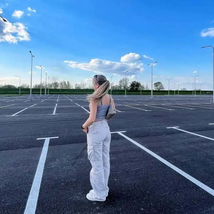 Woman standing in an empty parking lot wearing white cargo pants and a gray crop top, facing away from the camera.