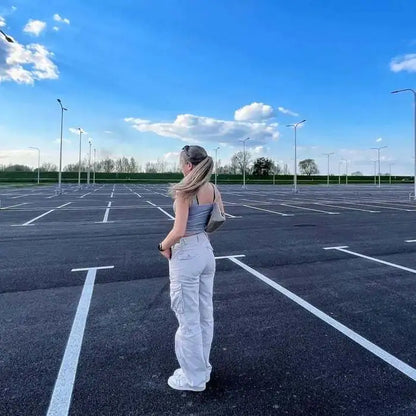 Woman standing in an empty parking lot wearing white cargo pants and a gray crop top, facing away from the camera.