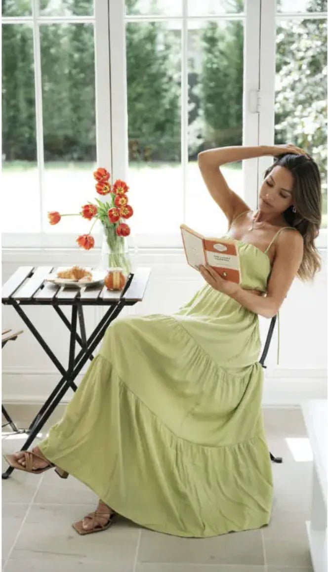 Woman sitting by a window reading a book while wearing a lime green open waist flared maxi dress with thin straps and tiered skirt.