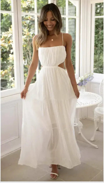 Woman wearing a white open waist flared maxi dress with thin straps and ruffle tiers, standing by a sunlit window.