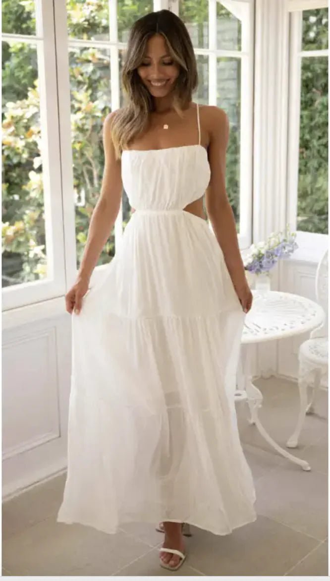 Woman wearing a white open waist flared maxi dress with thin straps and ruffle tiers, standing by a sunlit window.
