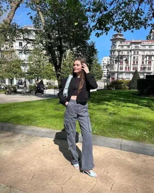 Stylish woman posing in a city park, wearing gray wide-leg cargo trousers with a black cropped jacket and blue heels.