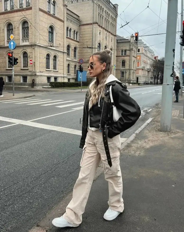Woman crossing a city street in beige cargo pants, layered with a black leather jacket and a white hoodie, paired with white sneakers.