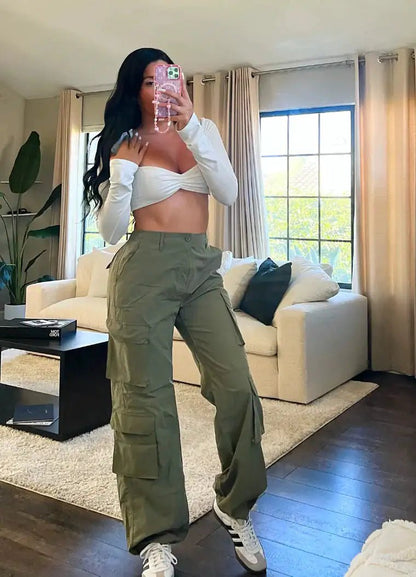 Woman taking a mirror selfie in olive green cargo trousers and a long-sleeve white off-shoulder crop top, standing in a stylish living room.