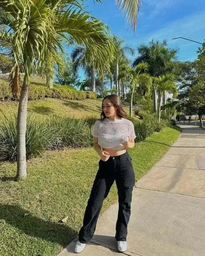 Woman standing outdoors on a sunny path, wearing high-waisted black utility trousers with a white mesh crop top and white sneakers.