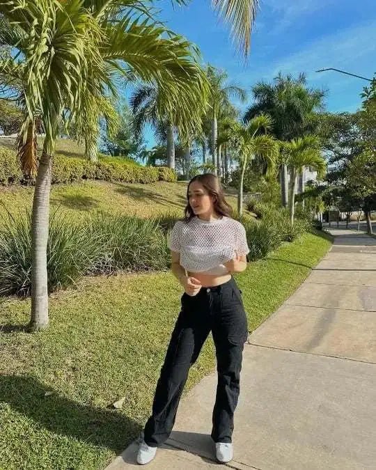 Woman standing outdoors on a sunny path, wearing high-waisted black utility trousers with a white mesh crop top and white sneakers.