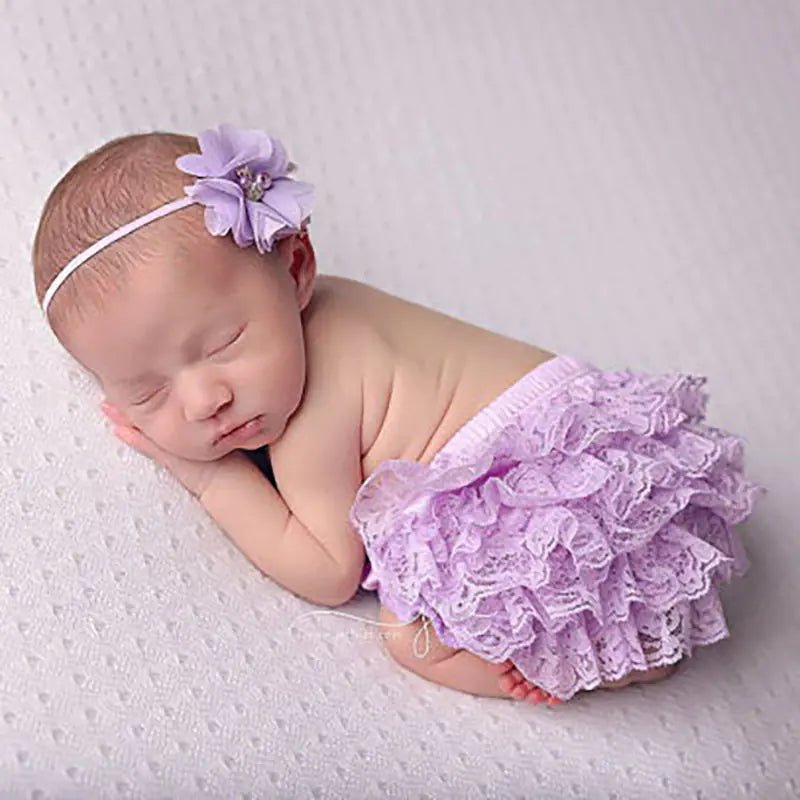 "Newborn baby sleeping in lavender lace bloomers with a matching flower headband on soft textured fabric"