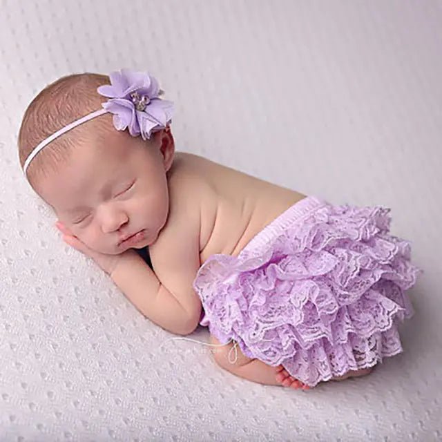 "Newborn baby sleeping in lavender lace bloomers with a matching flower headband on soft textured fabric"
