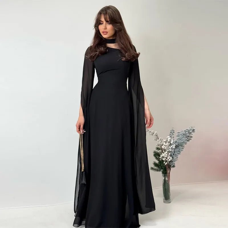 Woman dressed in a sleek black long dress with semi-sheer cape sleeves and a cutout neckline, accessorized with a small clutch.