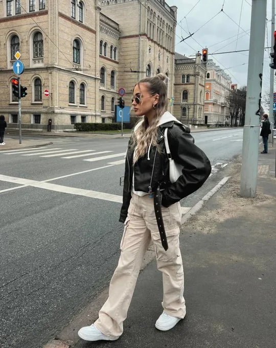 Woman crossing a city street in beige cargo pants, layered with a black leather jacket and a white hoodie, paired with white sneakers.