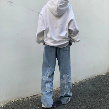 Back view of light blue wide-leg denim slacks with cross appliqué detail, worn with a white oversized hoodie, model walking on asphalt ground.