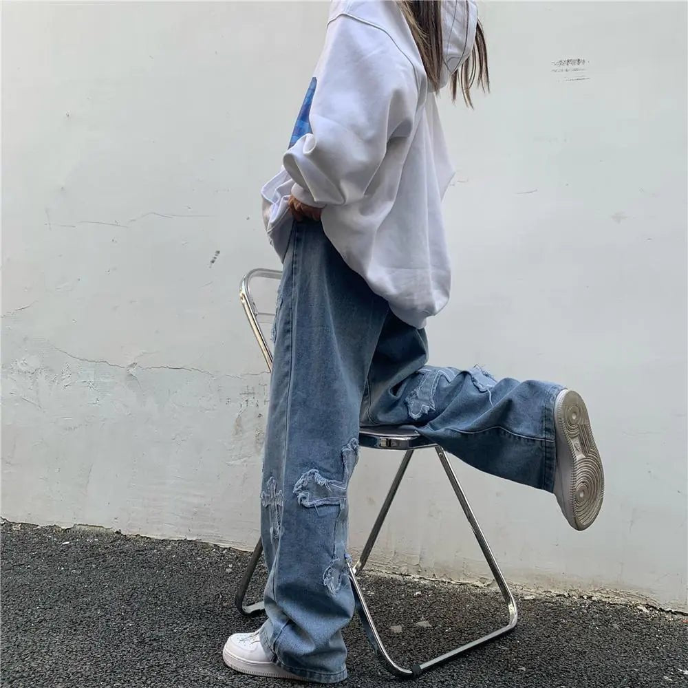 Side view of woman in relaxed-fit denim slacks with distressed patchwork and cross designs, paired with a white hoodie and white sneakers, leg raised on chair.