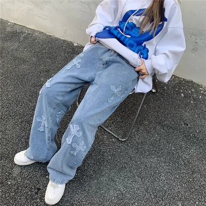 Close-up of high-waisted light blue denim slacks featuring cross appliqués, worn with a white oversized hoodie with blue print, model seated on a metal chair.