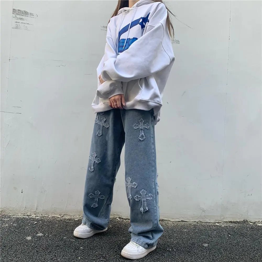 Woman wearing high-waisted denim slacks with distressed cross patches, paired with an oversized white hoodie and white sneakers, standing against a white wall.