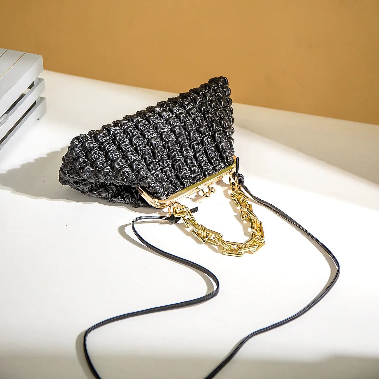 Black pleated handbag with chain detail displayed on a white seat.