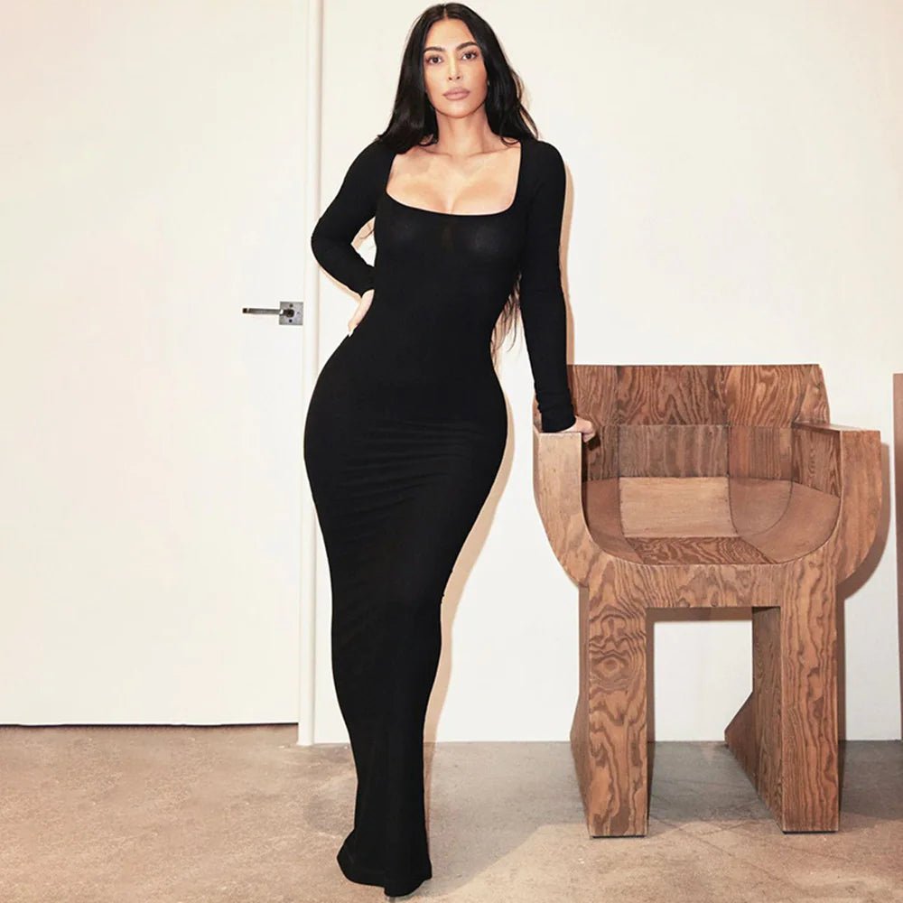 Woman in a black square neck bodycon dress leaning on a wooden chair in a modern interior setting.