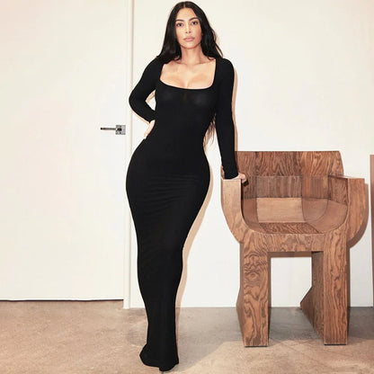Woman in a black square neck bodycon dress leaning on a wooden chair in a modern interior setting.