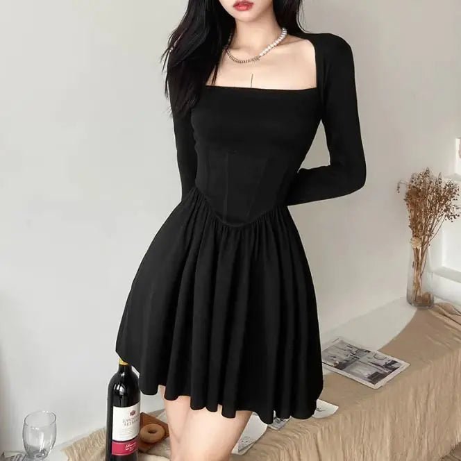 Front View: Woman wearing a black square neck mini dress with long sleeves, fitted bodice, and flared skirt, standing indoors beside a wine bottle.