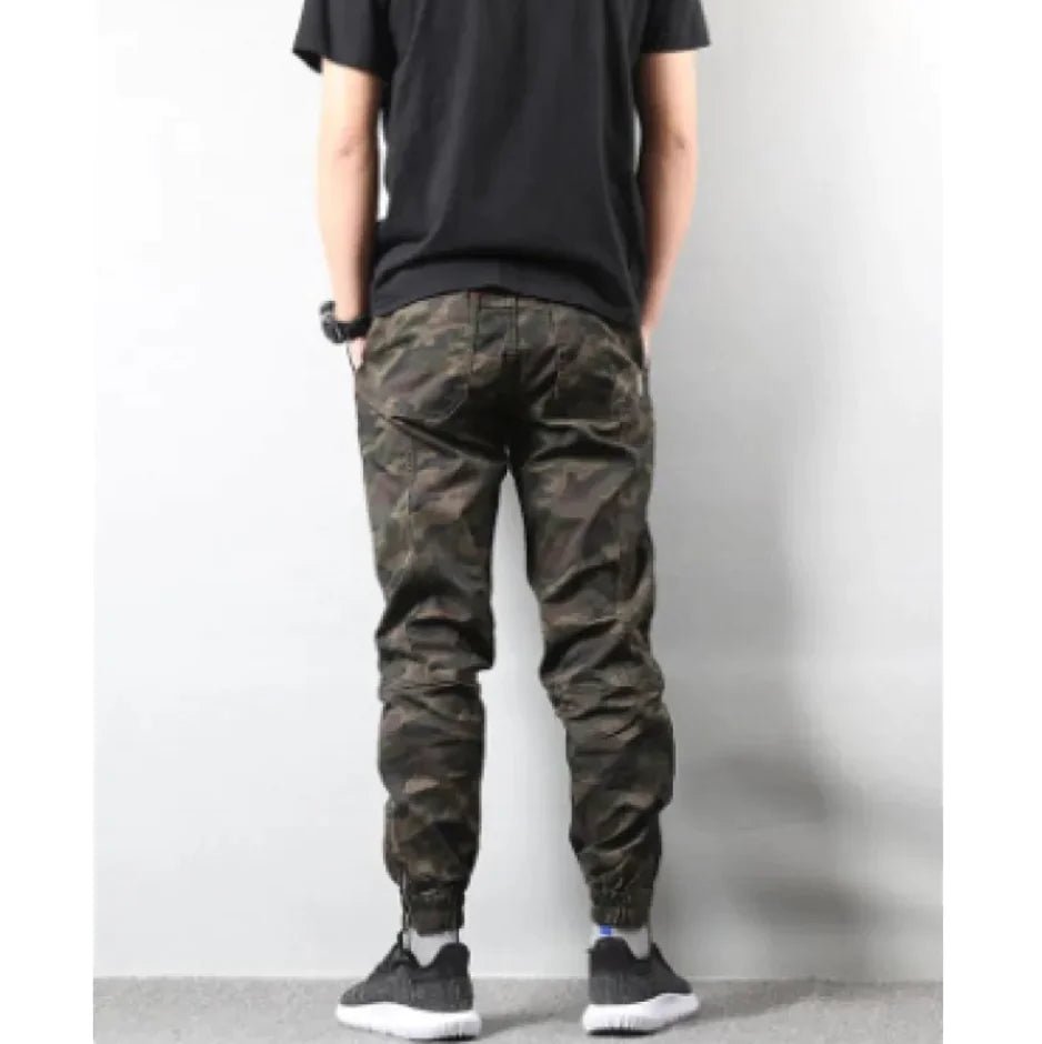 back view of a man dressed in streetwear-style camo pants and sneakers, posing casually against a light wall.
