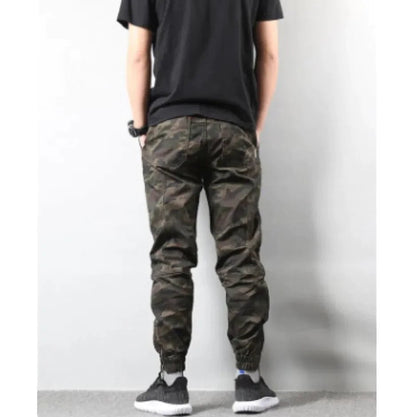 back view of a man dressed in streetwear-style camo pants and sneakers, posing casually against a light wall.