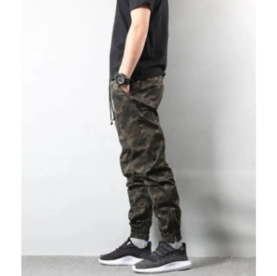 Side view of a man dressed in streetwear-style camo pants and sneakers, posing casually against a light wall.