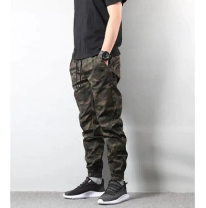 Side view of a man dressed in streetwear-style camo pants and sneakers, posing casually against a light wall.