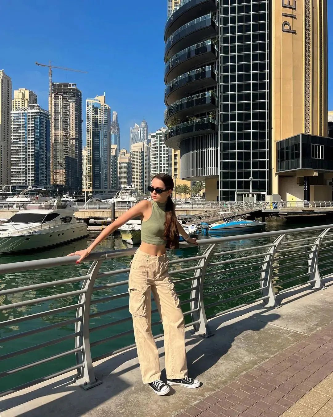 Woman wearing high-waisted beige cargo pants, standing on a city bridge with skyscrapers and yachts in the background.