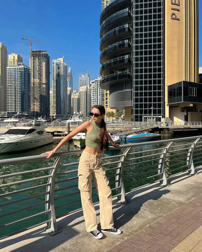 Woman wearing high-waisted beige cargo pants, standing on a city bridge with skyscrapers and yachts in the background.
