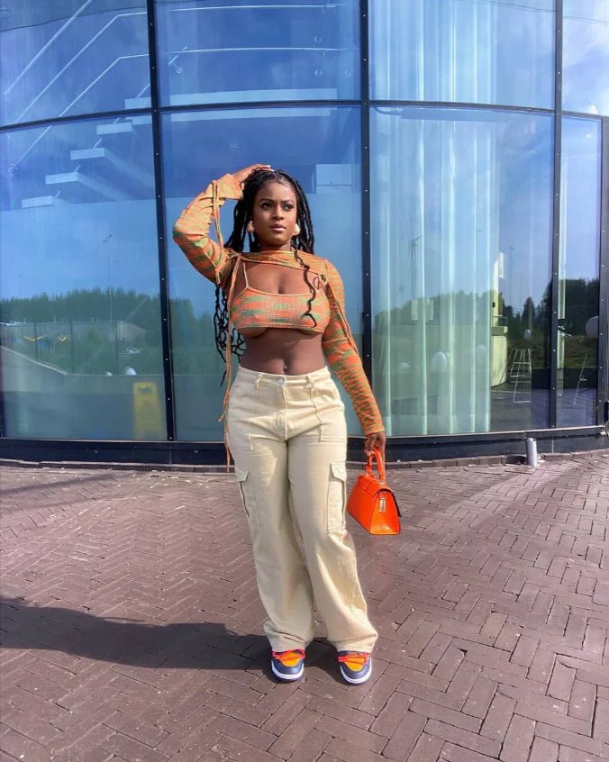 Woman wearing beige cargo pants with a multicolor crop top and orange handbag, standing confidently in front of a modern glass building.