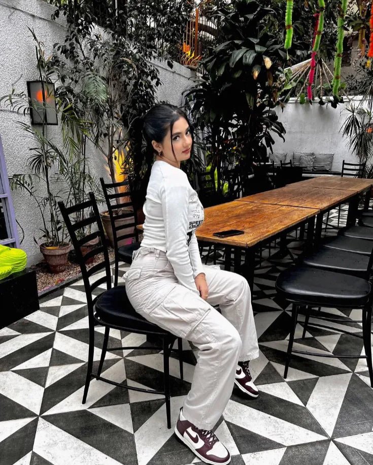 Woman sitting in a stylish indoor café wearing white cargo pants and a long-sleeve top, posing on a black chair.
