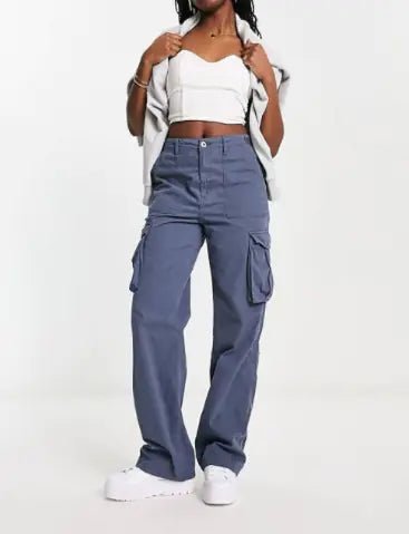Woman wearing blue cargo pants with a crop top and jacket draped over her shoulders, standing against a plain white background.